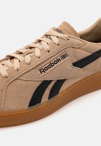 Sneaker in suede beige con dettagli neri, caratterizzato da un design con lacci, punta rotonda e una suola in gomma testurizzata. Logo impresso sul lato.