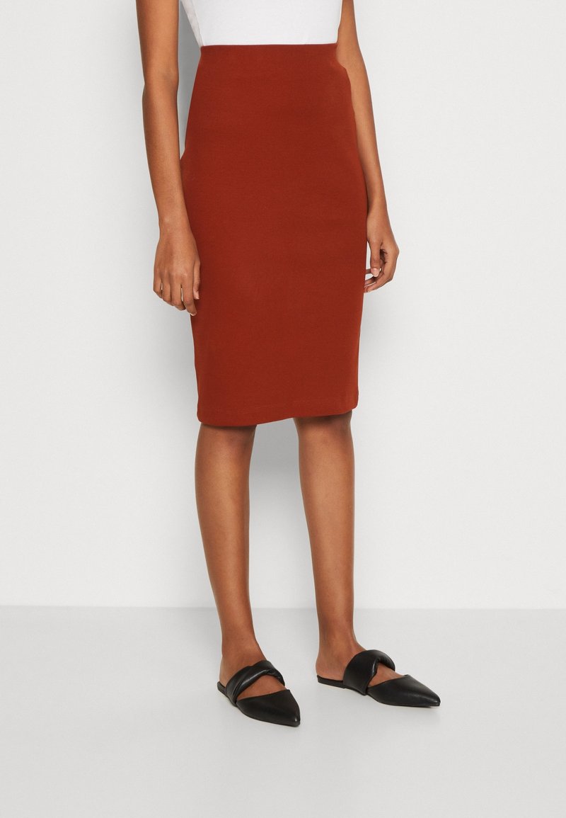 Femme portant une jupe ajustée couleur rouille mi-longue, un haut blanc et des chaussures plates noires pointues avec de larges brides.