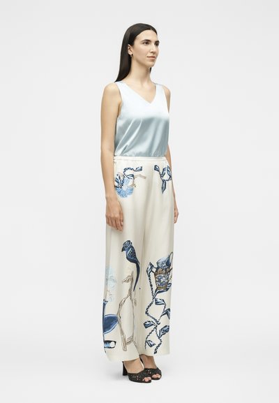 Femme portant un haut en satin bleu clair sans manches et un pantalon blanc taille haute à jambes larges avec des imprimés floraux et d'oiseaux bleus, debout avec des talons noirs.
