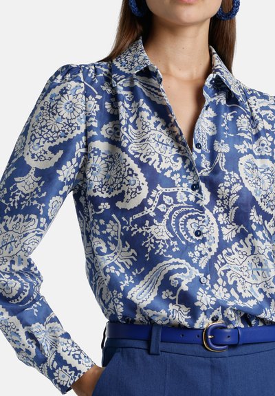 Blauw-wit bloemenpatroon blouse met een puntkraag en knopen, vervaardigd uit een gladde stof. Gecombineerd met een blauwe riem.