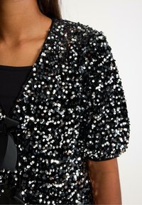 Černá blůza s flitry, texturovaným povrchem, kulatými flitry a nafouklými rukávy. Ozdobená černou stuhou.