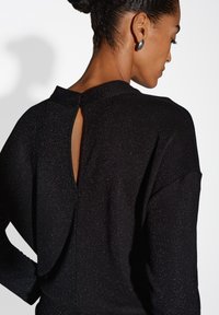 Donna con i capelli scuri raccolti in un bun, che indossa un top nero lucido con una piccola apertura a bottoncino sulla parte superiore della schiena e orecchini ovali argentati.