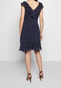 Navy blue polka dot dress with a flounce cap sleeve and a V-back. Features a ruffled hem and delicate texture. Paired with black strappy heels.