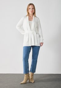 White knit cardigan with a waist tie, featuring a ribbed texture and two front pockets, paired with blue straight-leg jeans and beige heeled boots.