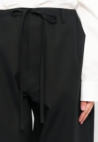 Sorte bukser med taljebånd, lavet af glat stof. Har sidelommer og en afslappet pasform, med en synlig krave fra en hvid skjorte.