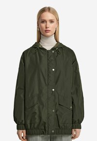 Olivenfärgad lätt jacka med huva, elastiska ärmmuddar och två framfickor. Har tryckknapps-stängning och dragsko i nederkant.