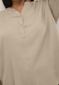 Beżowa lniana tunika z dekoltem henley, dwoma guzikami i krótkimi rękawami. Charakteryzuje się luźnym krojem i teksturowaną tkaniną.