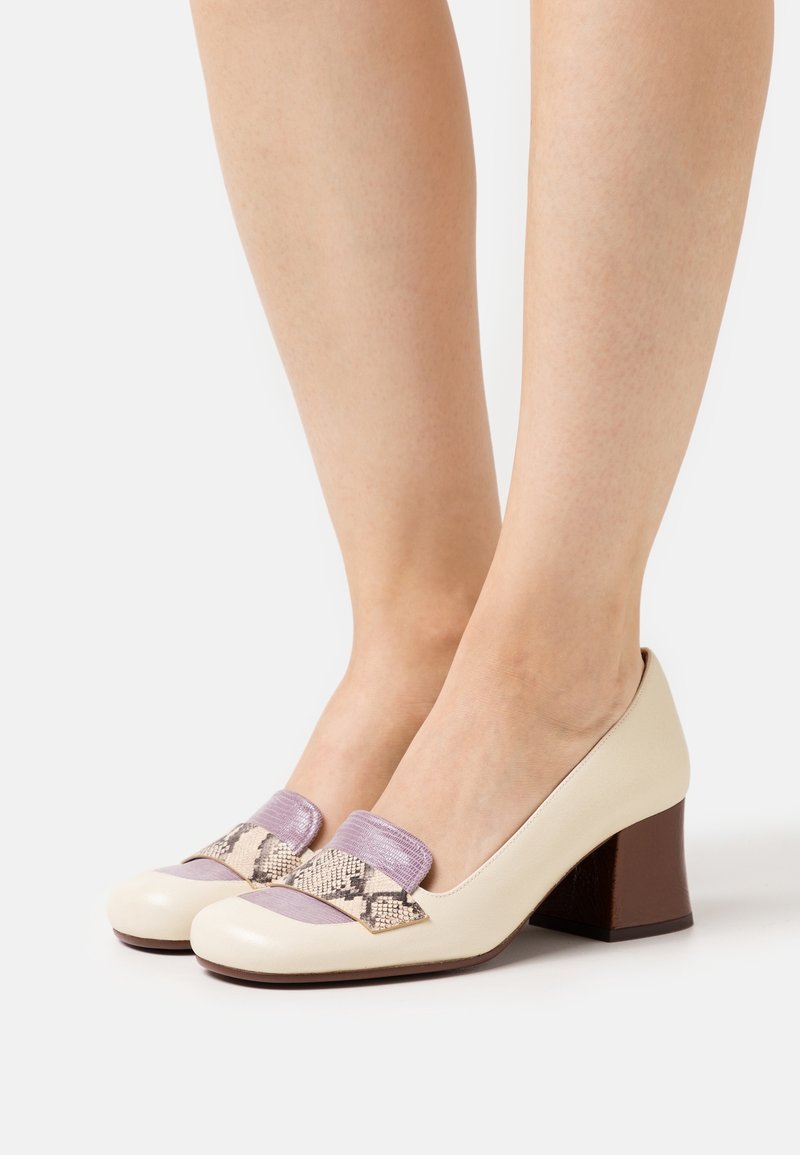 Femme portant des chaussures à talons blocs crème avec des détails en lavande et motif serpent sur le dessus, sur fond blanc.
