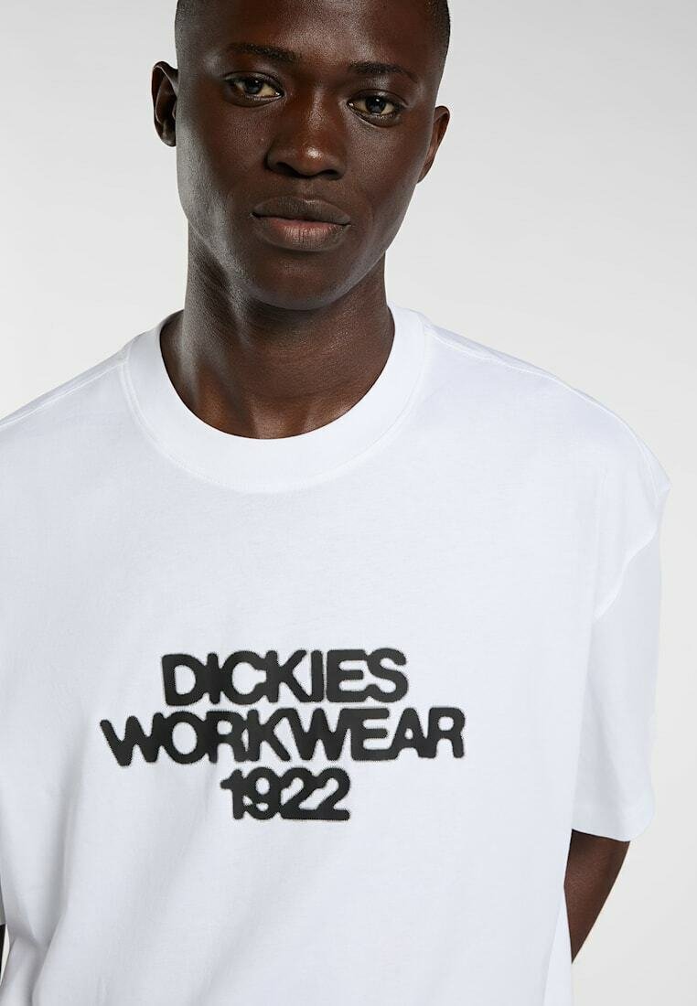 Junger schwarzer Mann trägt ein weißes T-Shirt mit dem in fettem schwarzen Text aufgedruckten Schriftzug "DICKIES WORKWEAR 1922" auf der Vorderseite.