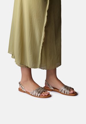 Pies con sandalias planas de tiras trenzadas metálicas combinadas con una falda plisada fluida amarillo-verde sobre un fondo blanco.