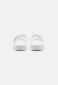 Bele sandale z gladko, mat teksturo, odprtim hrbtom in nastavljivim trakom za varen oprijem. Elegantna in minimalistična podoba.