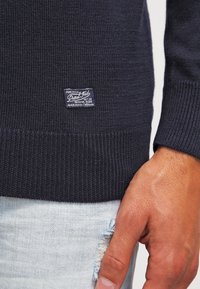 Pull en maille bleu marine avec ourlet et poignets côtelés. Comprend une étiquette cousue avec le logo. Main reposant sur un denim bleu clair usé.