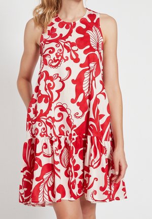 Biała sukienka bez rękawów z dużymi czerwonymi wzorami kwiatów i liści oraz falbaną na dole, noszona przez stojącą osobę.