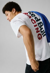 Camiseta de algodón blanca con el texto "Red Bull Racing" en negro y rojo. Una prenda azul está colgada sobre el hombro. Corte casual, mangas cortas.
