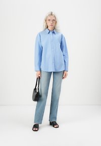 Camicia a righe blu e bianche con bottoni e maniche lunghe, abbinata a jeans a zampa d'elefante di un blu chiaro e sandali neri con tacco. Piccola borsa nera come accessorio.