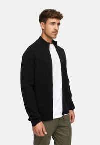Zwarte ritscardigan in een ribgebreide textuur, met een hoge kraag en een ontspannen pasvorm, gecombineerd met een witte T-shirt en olijfgroene broek.