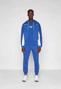 Lång man klädd i en blå och vit sportoverall med gula detaljer och vita sneakers, stående mot en enkel ljus bakgrund.