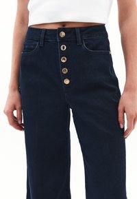 Granatowe dżinsowe spodnie z wysokim stanem, z pięcioma guzikami w rozporku, dwiema bocznymi kieszeniami i prostym krojem nogawki. Gładka faktura.