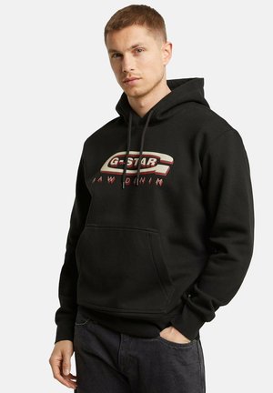 Schwarzer Hoodie mit einer vorderen Kängurutasche und einer regulierbaren Kapuze, ausgestattet mit einem auffälligen "G-Star Raw Denim"-Logo in kontrastierenden Farben.