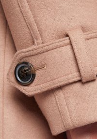 Beżowy wełniany płaszcz z szerokim mankietem, czarnym guzikiem i brązowymi akcentami, cechujący się miękką teksturą i dopasowanym krojem.