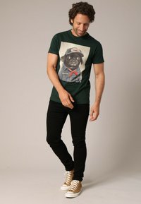 Groene T-shirt met een bedrukt ontwerp van een hond in een denimjasje en pet. Gecombineerd met zwarte broek en bruine hoge sneakers.