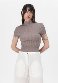 Brun ribbad kortärmad topp med en högre halsringning, figurnära design och en liten logodetalj, tillsammans med vita högmidjade byxor.