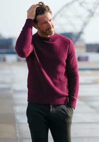 Burgunderroter Rippstrickpullover mit rundem Ausschnitt, kombiniert mit dunkelgrauen Hosen. Der Pullover zeichnet sich durch strukturierten Strick und eng anliegende Ärmel aus.
