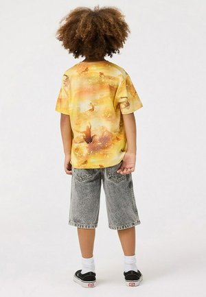 Maglietta grafica gialla con motivi astratti, abbinata a pantaloncini di jeans grigi chiari. Sneakers nere e calzini bianchi completano il look.