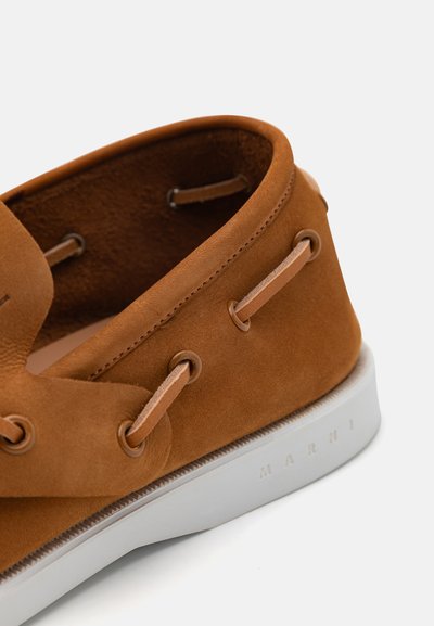 Brown suede boat shoe with white rubber sole, featuring contrast stitching and leather laces. Logo embossed on the side.