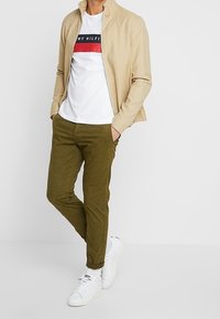 Beżowa kurtka zapinana na zamek na białej koszulce z czerwono-granatowym nadrukiem, oliwkowe spodnie i białe sneakersy. Luźny krój z gładkimi fakturami.