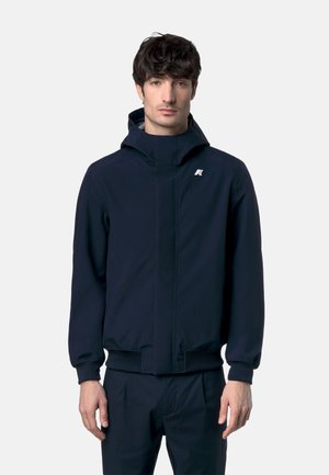 Uomo che indossa una giacca con cappuccio blu navy con un piccolo logo bianco sul petto, in piedi contro uno sfondo chiaro e neutro.