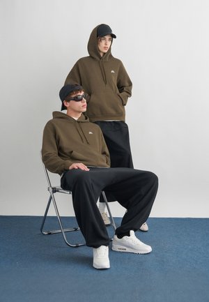 Jaunas vyras, sėdintis, ir moteris, stovinti, dėvi atitinkančius alyvinių atspalvių Nike džemperius su gaubtu, juodus apatinius drabužius ir kepures, pozuoja minimalistinėje studijos aplinkoje.