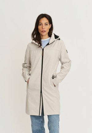 Frau trägt einen beigefarbenen, knielangen Kapuzenmantel mit Frontreißverschluss und Seitentaschen, kombiniert mit hellblauen Jeans und einem blauen Hemd.