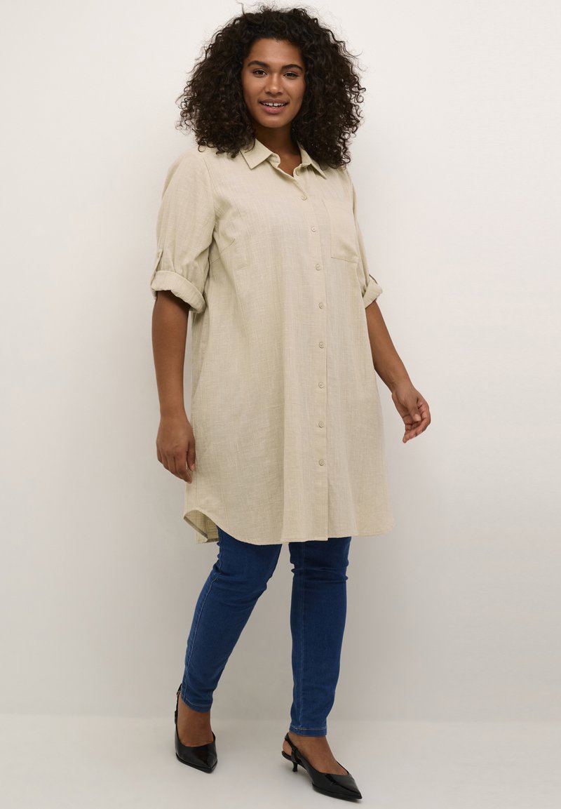 Robe chemise en lin beige avec un devant à boutons, des manches retroussées et une poche. Associée à un jean skinny bleu et des talons noirs.