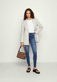 Mulher com cabelo encaracolado, vestindo um blusão bege, uma blusa branca, jeans skinny azuis, sandálias de plataforma pretas e segurando uma mala acolchoada castanha.