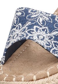 Chancla de textil azul con bordado floral blanco, plantilla de ante color marrón y suela de yute trenzado. Presenta un diseño de punta abierta y es fácil de poner.