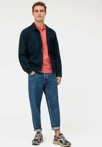 Donkerblauw overhemdjack, koraalroze poloshirt, lichtblauwe opgerolde spijkerbroek en sneakers in meerdere kleuren. Casual, ontspannen pasvorm met gelaagde stijl.