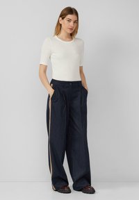 Witte geribde top met korte mouwen gecombineerd met marineblauwe broek met brede pinstripe en zijzakken, met een contrasterende beige streep aan de zijkanten van de broek.