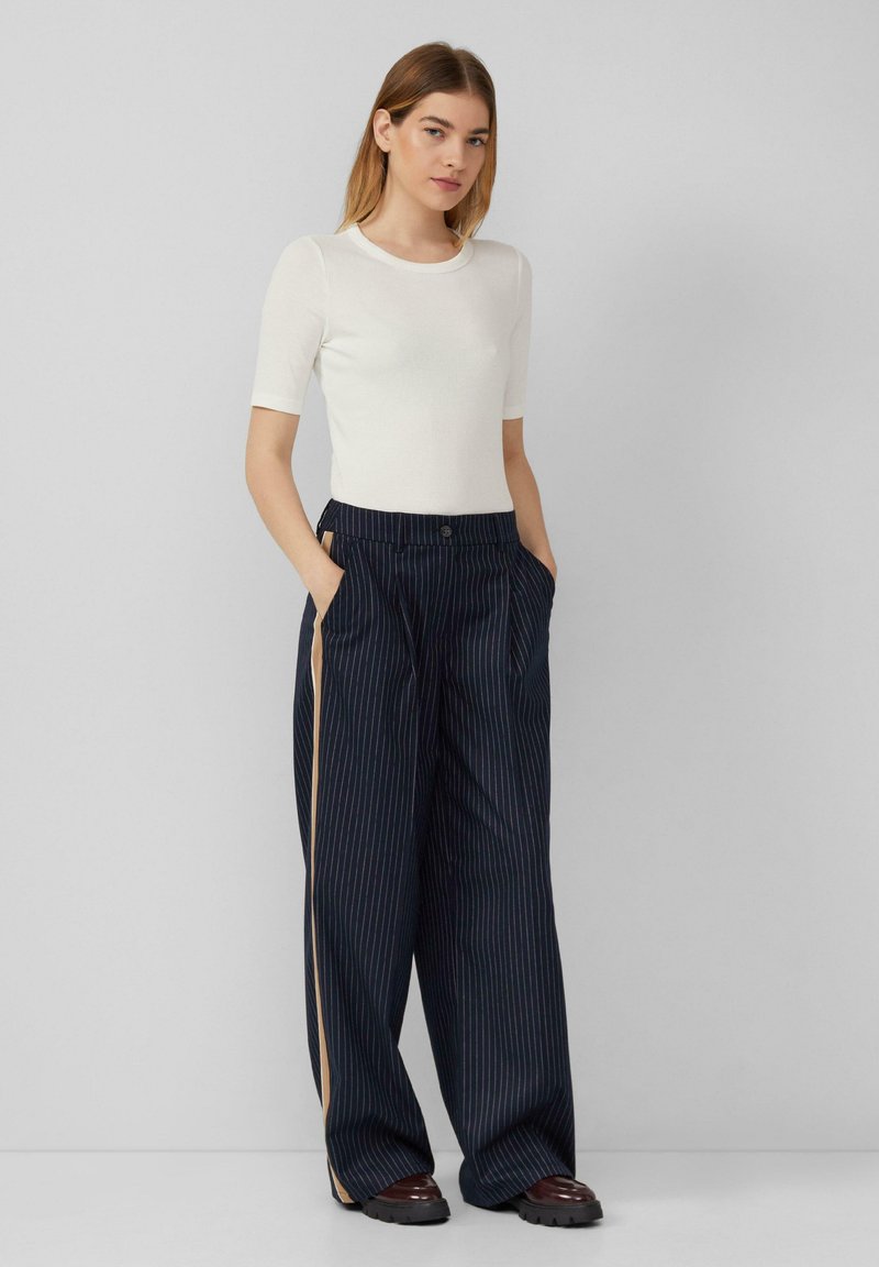 Witte geribde top met korte mouwen gecombineerd met marineblauwe broek met brede pinstripe en zijzakken, met een contrasterende beige streep aan de zijkanten van de broek.