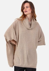 Pull-over beige oversize avec un col roulé côtelé, des manches chauve-souris courtes, une texture douce et une silhouette décontractée. Stylisé avec un jean foncé.