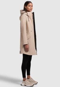 Beige vattentät jacka med huva, frontdragkedja och sidofickor, bärs över svarta tights och ljusa sneakers, visad mot en grå bakgrund.