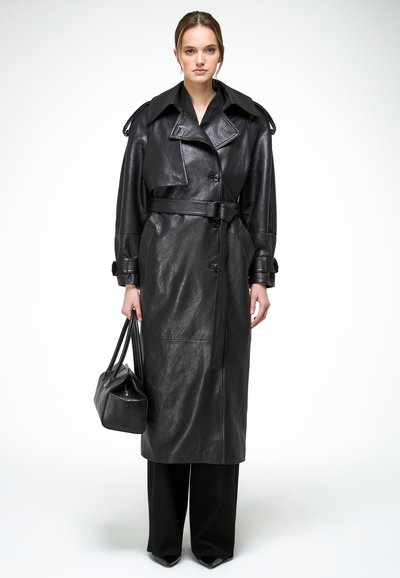 Vrouw draagt een lange zwarte leren trenchcoat met riem, houdt een zwarte handtas vast en staat voor een effen witte achtergrond.