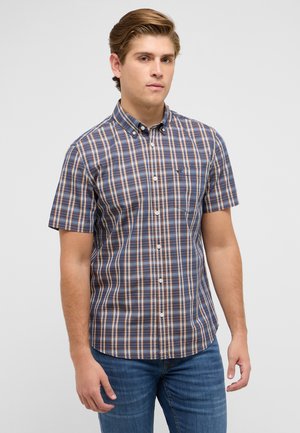 Junger Mann steht, trägt ein blau-orange kariertes kurzärmliges Hemd und blaue Jeans vor einem einfarbigen Hintergrund.