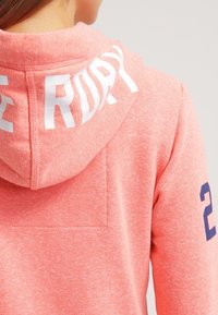 Persona che indossa una felpa con cappuccio di colore rosa corallo, con la scritta bianca "THE ROCKY" sul cappuccio e il numero blu navy "23" sulla manica.