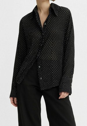Femme portant une chemise noire à boutons avec de petits motifs en tirets blancs et un pantalon noir ample, se tenant devant un fond uni.