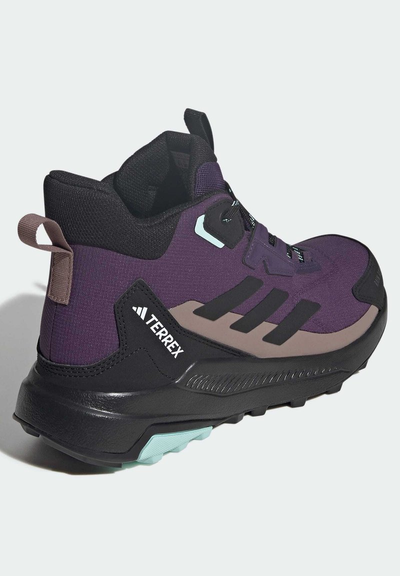 Zapato de senderismo púrpura con acentos negros, parte superior texturizada y suela de goma resistente. Presenta un logo y una lengüeta en la parte trasera para facilitar su uso.