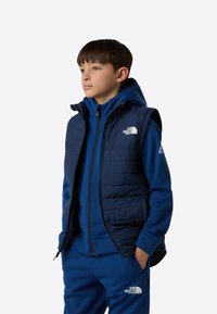 Jachetă bleumarin cu glugă din fleece, cu fermoar, purtată sub o vestă fără mâneci matlasată, tot bleumarin. Ambele au logo-ul The North Face. Material texturat vizibil.