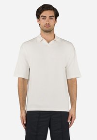 Short-sleeve, cream polo shirt made from soft fabric. Features a V-neck collar and relaxed fit, with minimal stitching detail.