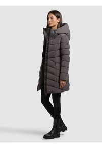 Parka gris acolchada con capucha, diseño acolchado, cremallera frontal y detalles en los puños. Combinada con leggings negros y botas negras gruesas.