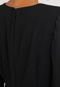 Schwarzes Kleid mit hohem Ausschnitt, dezentem Reißverschluss auf der Rückseite und plissierten, strukturierten Ärmeln. Glattes Material mit weichem Finish.